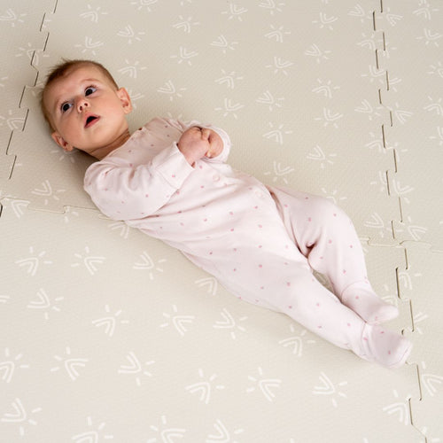 A baby in a light pink onesie lies on a TP Toys TP Active-Tots Safety Play Mat, featuring a patterned, non-toxic foam surface, looking up with hands clasped.