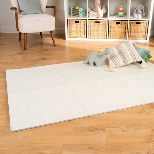 The TP Toys TP Active-Tots Safety Play Mats feature a subtle pattern and non-toxic foam, creating a cozy toddler play area on the wooden floor beside a plush dinosaur, cushion, and shelving unit with baskets and toys.