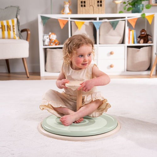 A young child sits cross-legged on the TP Active-Tots Pikler Style Wooden Spinning Seat - FSC® certified by TP Toys, enjoying sensory play in a living room with shelves, toys, and decorative bunting in the background.