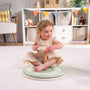 A young child sits cross-legged on the TP Active-Tots Pikler Style Wooden Spinning Seat - FSC® certified by TP Toys, enjoying sensory play in a living room with shelves, toys, and decorative bunting in the background.
