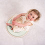 A young child in a cream dress sits on the TP Active-Tots Pikler Style Wooden Spinning Seat by TP Toys (FSC® certified), a round, mint green Montessori-inspired toddler toy, smiling and looking up while on a light-colored carpet.