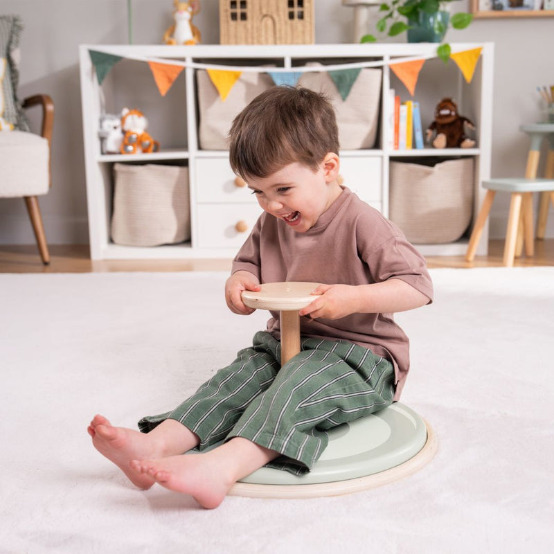 A young child sits and spins on the TP Active-Tots Pikler Style Wooden Spinning Seat by TP Toys, an FSC® certified Montessori toddler toy, in a playroom with shelves, books, and toys in the background.