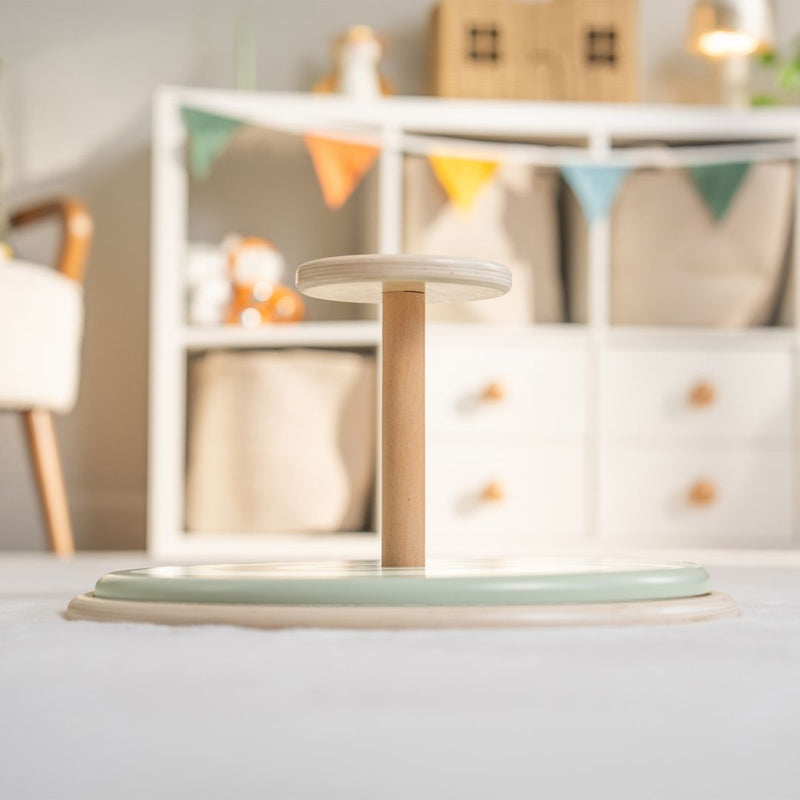The TP Toys Active-Tots Pikler Style Wooden Spinning Seat—crafted from FSC® certified wood—stands on a light-colored floor in a children’s playroom, surrounded by shelves and colorful pennant bunting in the background.
