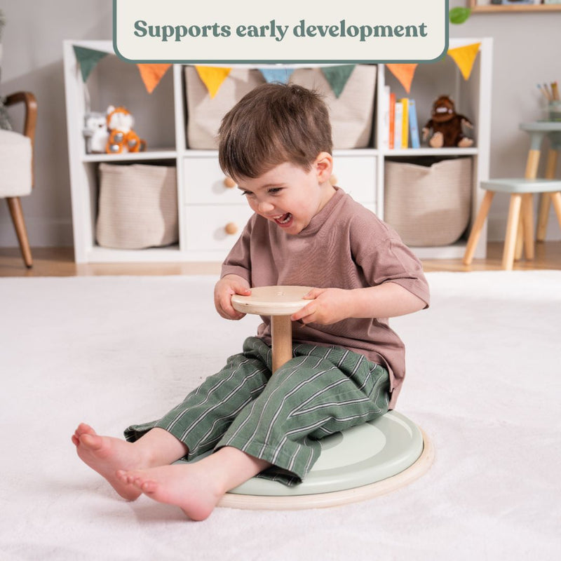 A young child sits on the TP Active-Tots Pikler Style Wooden Spinning Seat by TP Toys in a playroom, smiling and gripping the handle, with toys and storage baskets in the background. This FSC® certified sensory toy supports early development. Text above reads, Supports early development.