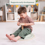 A young child sits on the TP Active-Tots Pikler Style Wooden Spinning Seat by TP Toys in a playroom, smiling and gripping the handle, with toys and storage baskets in the background. This FSC® certified sensory toy supports early development. Text above reads, Supports early development.