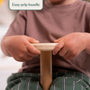 A child grips the FSC® certified wooden handle of the TP Active-Tots Pikler Style Wooden Spinning Seat by TP Toys with both hands, wearing a brown shirt and striped green pants. Text reads: Easy grip handle—perfect for sensory play and Montessori toddler toy activities.