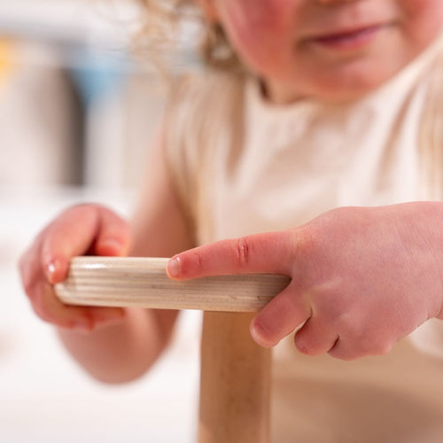 A young child grips the smooth wooden bar of the TP Toys Active-Tots Pikler Style Wooden Spinning Seat—FSC® certified—with both hands, engaging in sensory play. The child wears a light-colored sleeveless shirt; only the lower part of their face is visible.