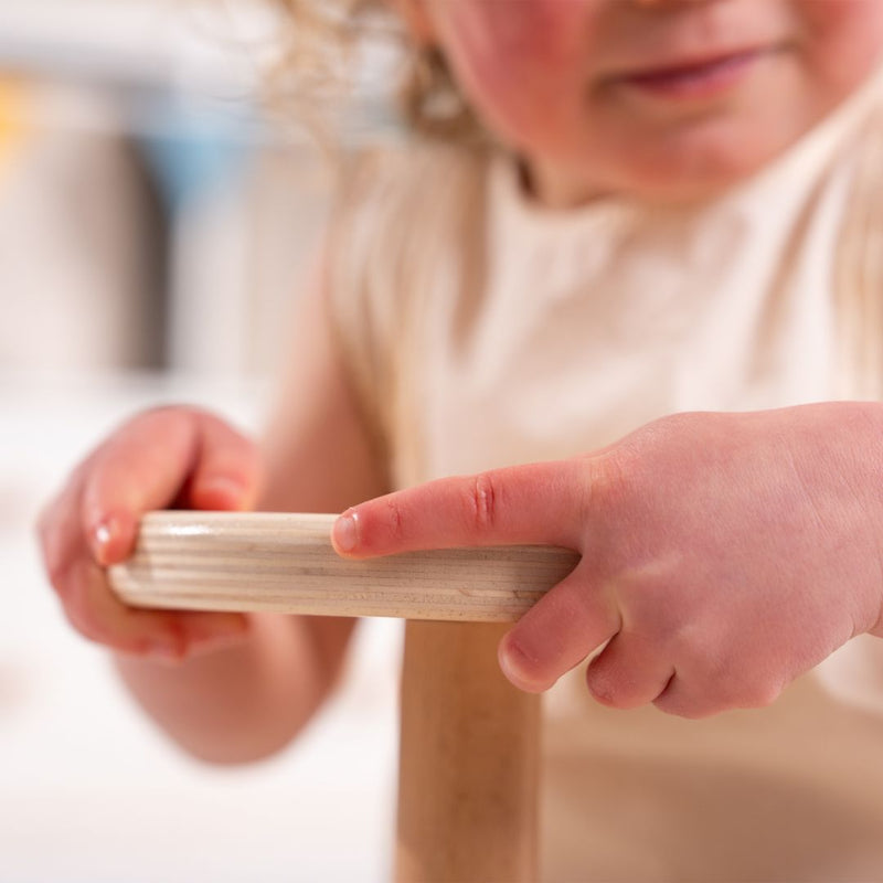 A young child grips the smooth wooden bar of the TP Toys Active-Tots Pikler Style Wooden Spinning Seat—FSC® certified—with both hands, engaging in sensory play. The child wears a light-colored sleeveless shirt; only the lower part of their face is visible.