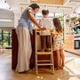 A young child stands on a TP Active-Tots Pikler Style Wooden Helper Tower at a kitchen island while an adult and another child stand beside them.