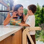 A woman playfully pretends to be a tiger while reading to a young boy standing in a TP Active-Tots Pikler Style Wooden Helper Tower at the kitchen counter.