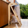 A child stands on a TP Active-Tots Pikler Style Wooden Helper Tower at a kitchen counter next to an adult.