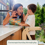 A child stands on a TP Active-Tots Pikler Style Wooden Helper Tower at a kitchen counter, reading a tiger book with an adult making a playful expression, under the text: Encourages learning through play.