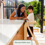 A child stands in a TP Active-Tots Pikler Style Wooden Helper Tower - FSC® certified at a kitchen counter, reading a book with a smiling woman.