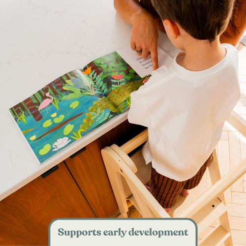 A child stands on a TP Active-Tots Pikler Style Wooden Helper Tower at a counter, reading a picture book with an adult.