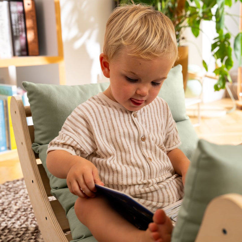 A toddler with blonde hair sits on the TP Toys TP Active-Tots Sway Wooden Rocker with Cushion (FSC® certified), intently looking at a book in a sunlit room decorated with plants and bookshelves.
