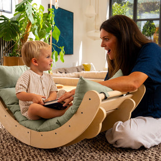 A woman sits on the floor, smiling at a young child seated in the TP Toys TP Active-Tots Sway Wooden Rocker with Cushion (FSC® certified), surrounded by plants and natural light in a cozy living room.