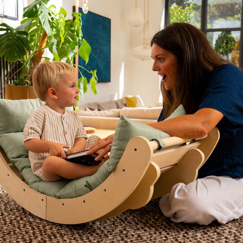 A woman sits on the floor, smiling at a young child seated in the TP Toys TP Active-Tots Sway Wooden Rocker with Cushion (FSC® certified), surrounded by plants and natural light in a cozy living room.