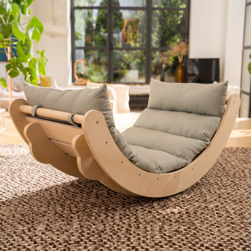 The TP Toys Active-Tots Sway Wooden Rocker with Cushion—crafted from FSC® certified wood—sits atop a textured rug in a sunlit living room, surrounded by large windows and indoor plants, making it an ideal Montessori-inspired toddler rocker that combines style and comfort.