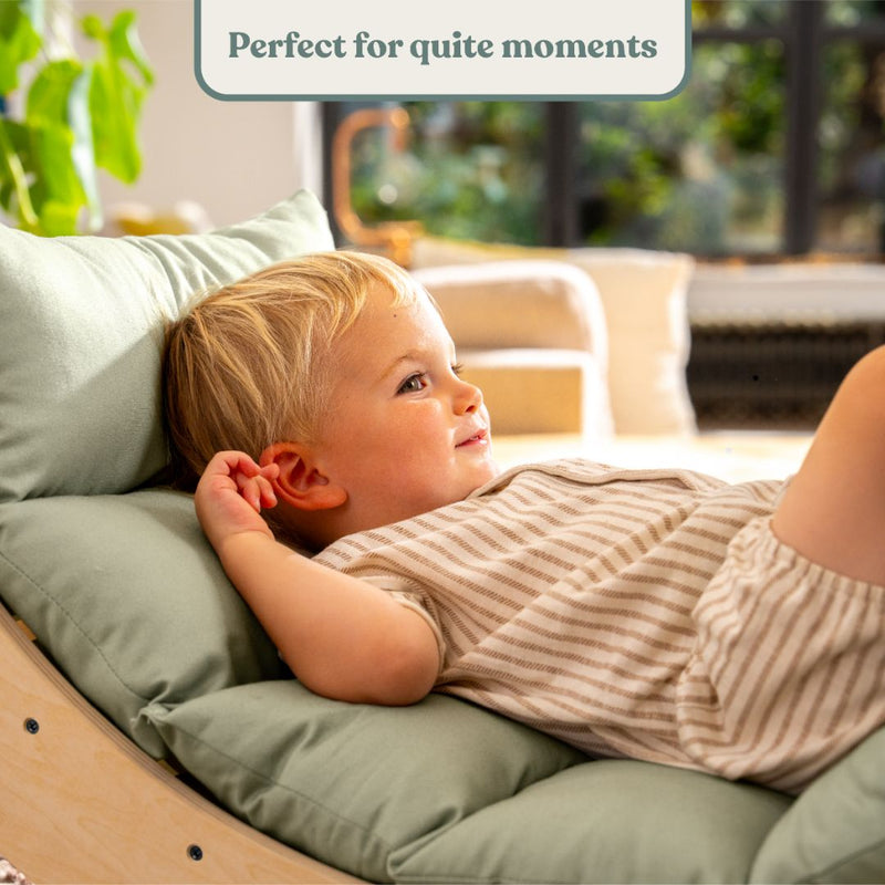 A young child reclines on the TP Toys Active-Tots Sway Wooden Rocker with Cushion (FSC® certified), smiling and looking relaxed indoors. Text above reads, Perfect for quiet moments.