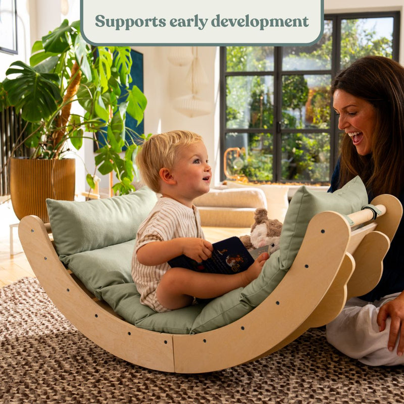A young child sits in the TP Toys Active-Tots Sway Wooden Rocker with Cushion (FSC® certified), holding a book and smiling at an adult woman. Text above reads, Supports early development. They are in a bright, cozy room.