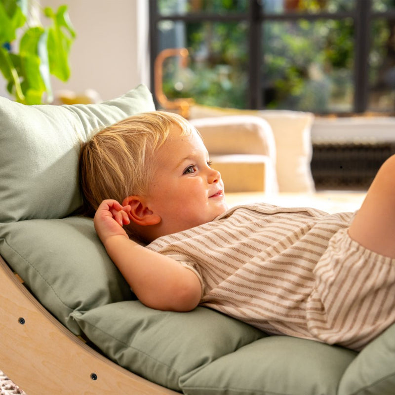 A young child with blonde hair reclines on the TP Active-Tots Sway Wooden Rocker with Cushion by TP Toys, featuring a light green padded seat, gazing off to the side indoors with a relaxed expression.