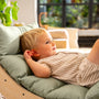 A young child with blonde hair reclines on the TP Active-Tots Sway Wooden Rocker with Cushion by TP Toys, featuring a light green padded seat, gazing off to the side indoors with a relaxed expression.
