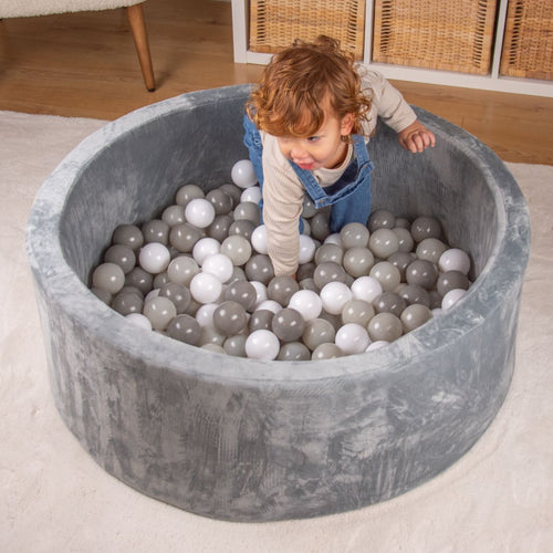 A young child in overalls leans into a round, gray Active Tots Foam Ball Pit with 100 Playballs, which is filled with white, gray, and clear balls for sensory play.
