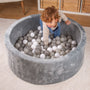 A young child in overalls leans into a round, gray Active Tots Foam Ball Pit with 100 Playballs, which is filled with white, gray, and clear balls for sensory play.
