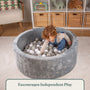 A young child enjoys sensory play alone in the TP Toys Active Tots Foam Ball Pit, which is filled with 100 white and grey playballs, in a cozy, organized playroom.