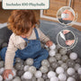 A toddler dressed in blue overalls sits in the TP Toys Active Tots Foam Ball Pit with 100 Playballs, surrounded by soft velvet fabric and a mix of white, gray, and beige air-filled balls for a delightful sensory play experience. Insets showcase close-ups of the playballs and the plush velvet texture.