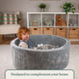 A young child sits in the TP Toys Active Tots Foam Ball Pit with 100 Playballs, featuring a gray velvet finish and filled with neutral-colored playballs, engaging in sensory play with shelves and toys visible in the background.