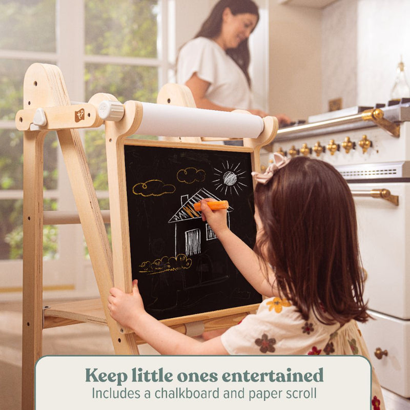 A young child draws on the TP Toys TP Active-Tots Pikler Style Wooden Easel Play Accessory while an adult works in the kitchen. Keeps little ones entertained with a chalkboard and paper scroll. FSC® certified.