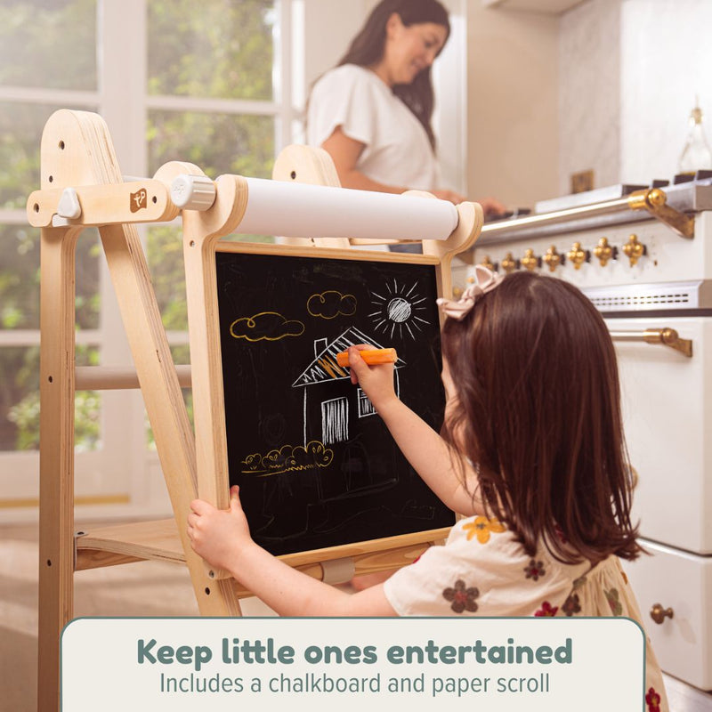A young girl draws on the chalkboard easel of the TP Active-Tots Pikler Style Wooden Folding Helper Tower by TP Toys while an adult looks on. Keep little ones engaged with this adjustable, FSC® certified tower that includes a chalkboard and paper scroll.