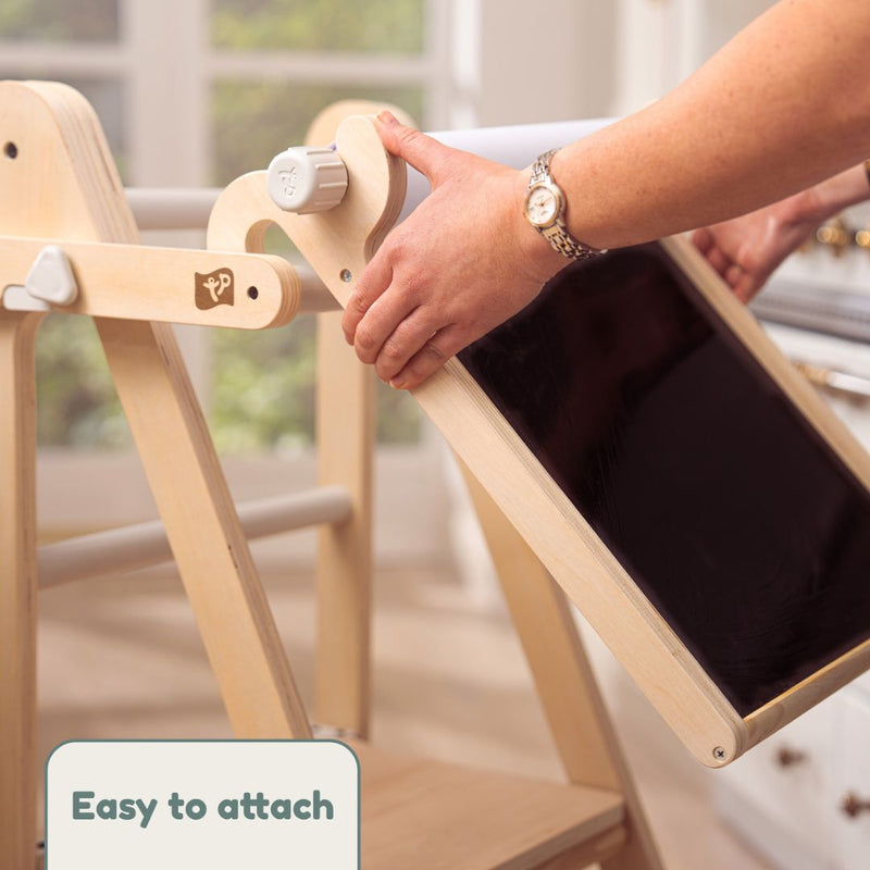 In a bright room, someone attaches the TP Toys TP Active-Tots Pikler Style Wooden Folding Helper Tower With Play Easel – FSC® certified to an easel. A label in the corner reads "Easy to attach.