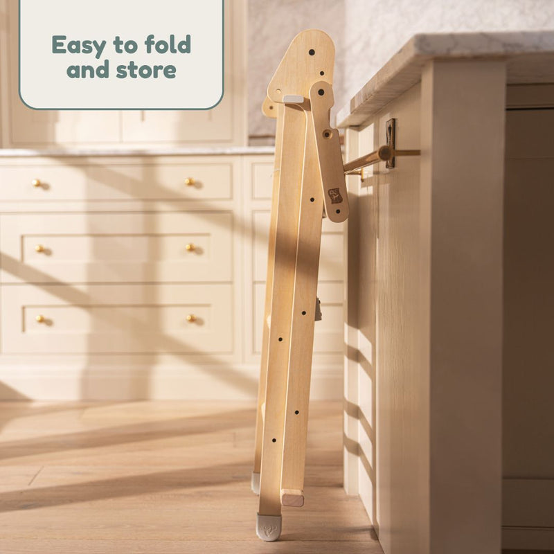 The TP Active-Tots Pikler Style Wooden Folding Helper Tower With Play Easel by TP Toys stores flat against a cabinet, featuring a sign that says "Easy to fold and store.