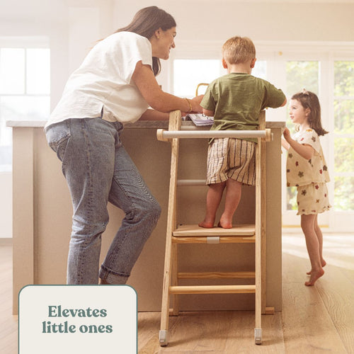 A woman stands by a kitchen island with a young boy on the TP Toys Active-Tots Pikler Style Wooden Folding Helper Tower – FSC® certified, and a girl nearby. Text reads, Elevates little ones.