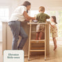 A woman stands by a kitchen island with a young boy on the TP Toys Active-Tots Pikler Style Wooden Folding Helper Tower – FSC® certified, and a girl nearby. Text reads, Elevates little ones.