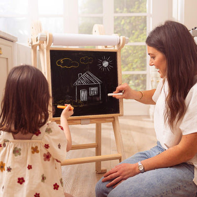 An adult and child use the TP Toys TP Active-Tots Pikler Style Wooden Folding Helper Tower With Play Easel & Play Mats to draw a house, sun, and clouds on the easel in a bright room, reaching comfortably with this FSC® certified product.