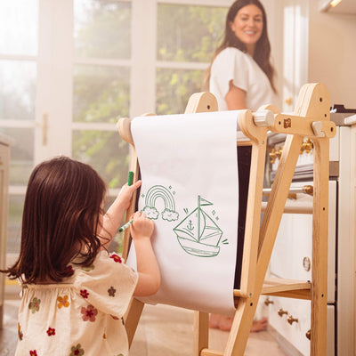 A young child stands on the TP Toys Active-Tots Pikler Style Wooden Folding Helper Tower with Play Easel, drawing on a paper roll, while an adult woman smiles in the background in a bright, cheerful room.
