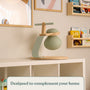 A modern green and wooden lamp sits on a white shelf in a children’s room filled with books and art, creating a cozy play space complemented by the TP Toys TP Active-Tots Pikler Style Toddler Bouncer—FSC® certified for gross motor skill development.