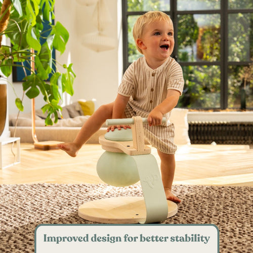 A toddler in a striped romper sits on the TP Active-Tots Pikler Style Toddler Bouncer - FSC® certified by TP Toys, featuring mint green accents, in a bright, modern living room. Text reads: Improved design for better stability.