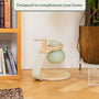 A light green modern kettle with a wooden handle sits on a matching circular wooden stand beside a bookshelf, the TP Toys TP Active-Tots Pikler Style Toddler Bouncer - FSC® certified, and a plant in a cozy living space.