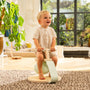 A young child sits and smiles on the TP Toys TP Active-Tots Pikler Style Toddler Bouncer – FSC® certified, in a sunlit living room with large windows, a rug, and indoor plants.