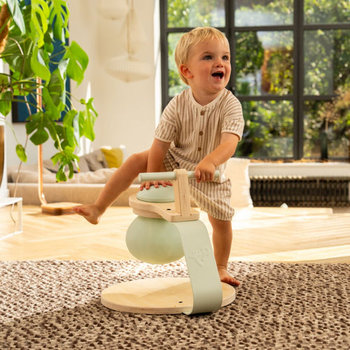 A toddler in striped pajamas balances on one foot atop the TP Active-Tots Pikler Style Toddler Bouncer by TP Toys (FSC® certified) in a sunlit living room with a large window and an indoor plant.