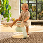 A toddler in striped pajamas balances on one foot atop the TP Active-Tots Pikler Style Toddler Bouncer by TP Toys (FSC® certified) in a sunlit living room with a large window and an indoor plant.
