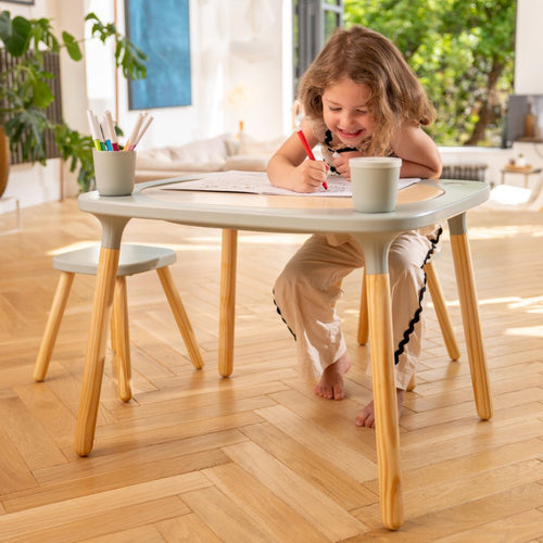 A young girl draws with colored pens on paper at the TP Toys Active-Tots Pikler Style 4 in 1 Wooden Play Table with two play stools (FSC® certified) in a bright, sunlit room with wooden floors and plants.