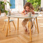 A young girl draws with colored pens on paper at the TP Toys Active-Tots Pikler Style 4 in 1 Wooden Play Table with two play stools (FSC® certified) in a bright, sunlit room with wooden floors and plants.