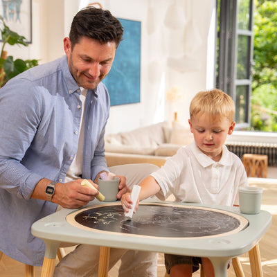 An adult and a young boy draw on the TP Toys Active-Tots Pikler Style 4 in 1 Wooden Play Table (FSC® certified) in a bright living room, with mugs placed on the table.