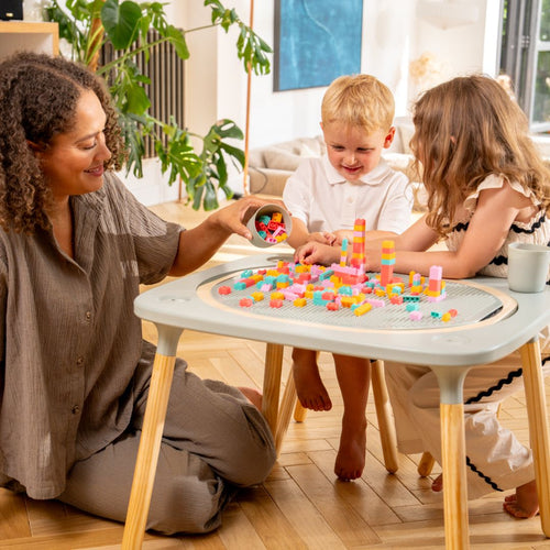 An adult and two kids use the TP Toys Active-Tots Pikler Style 4 in 1 Wooden Play Table, building with colorful blocks in a bright, modern living room. The FSC® certified table is perfect for Montessori-inspired activities.