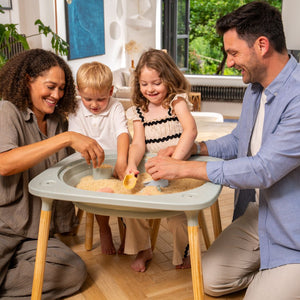 Two adults and two children play with sand and toy scoops at the TP Toys Active-Tots Pikler Style 4 in 1 Wooden Play Table (FSC® certified) in a bright, modern living room.